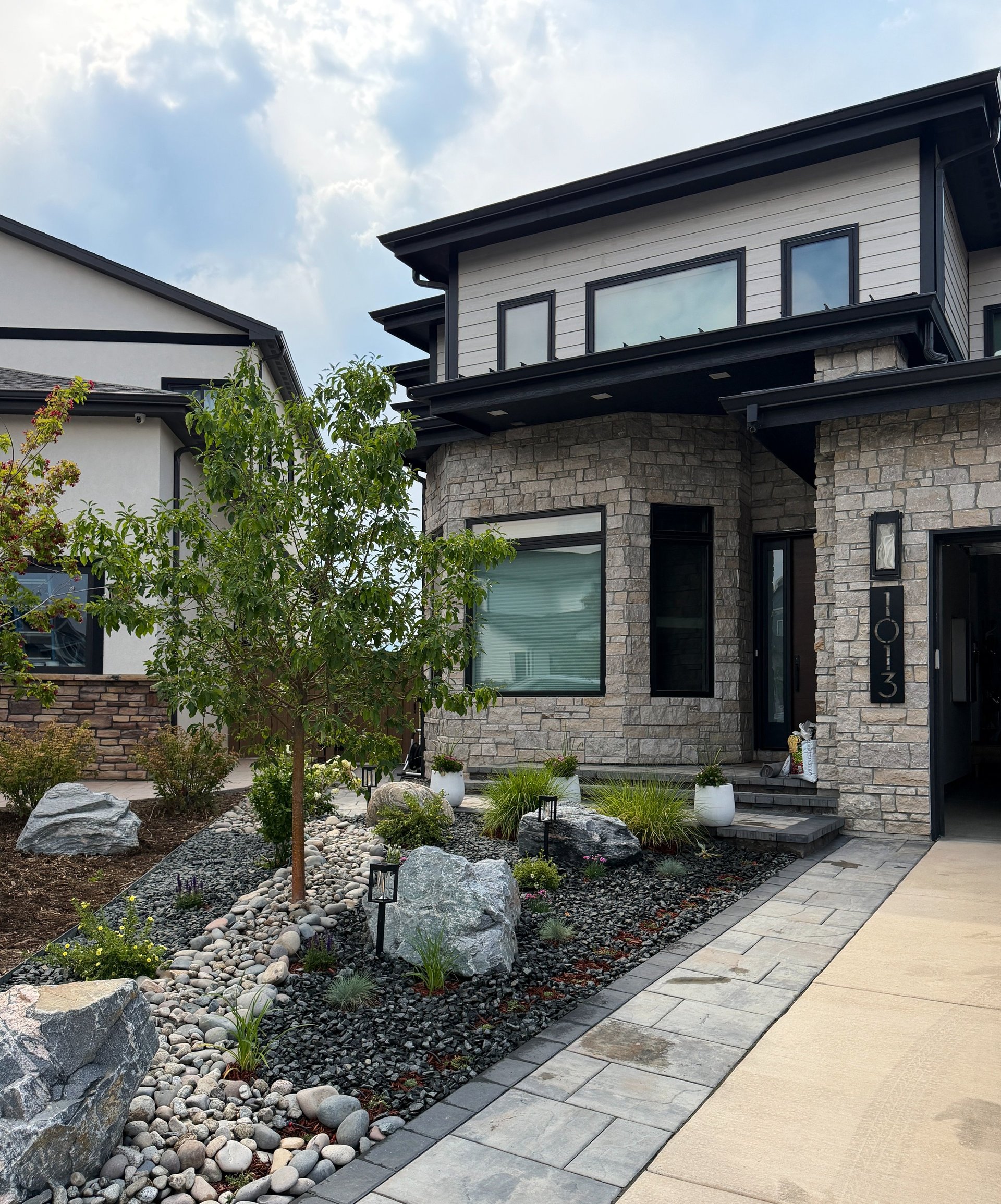 Modern two-story home with black and white exterior, stone base, large windows, and contemporary landscaping with young trees and gravel