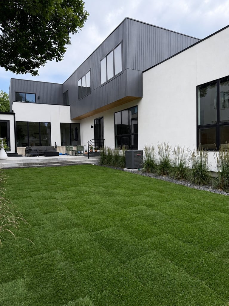 Modern two-story house with black metal siding and white walls, featuring large windows, patio area, and manicured lawn in front.