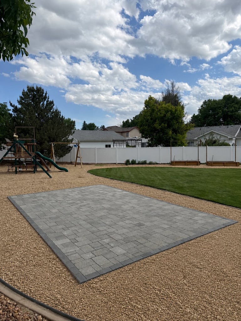 Large residential backyard with paver patio, playground equipment, green lawn, white fence, and trees under partly cloudy sky