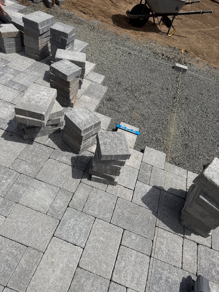 Construction site with gray concrete pavers laid out in rows, stacked blocks, and a wheelbarrow in the background on dirt ground