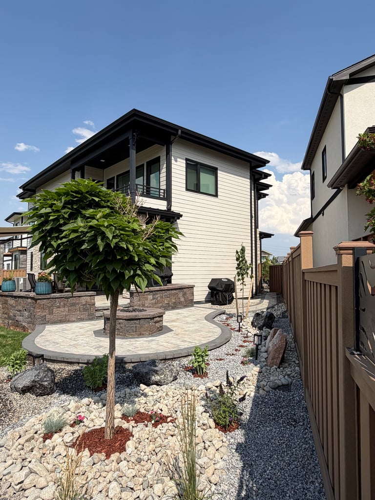 Modern white two-story house with black roof trim, circular patio with young tree, rock landscaping, and decorative fencing on a clear day
