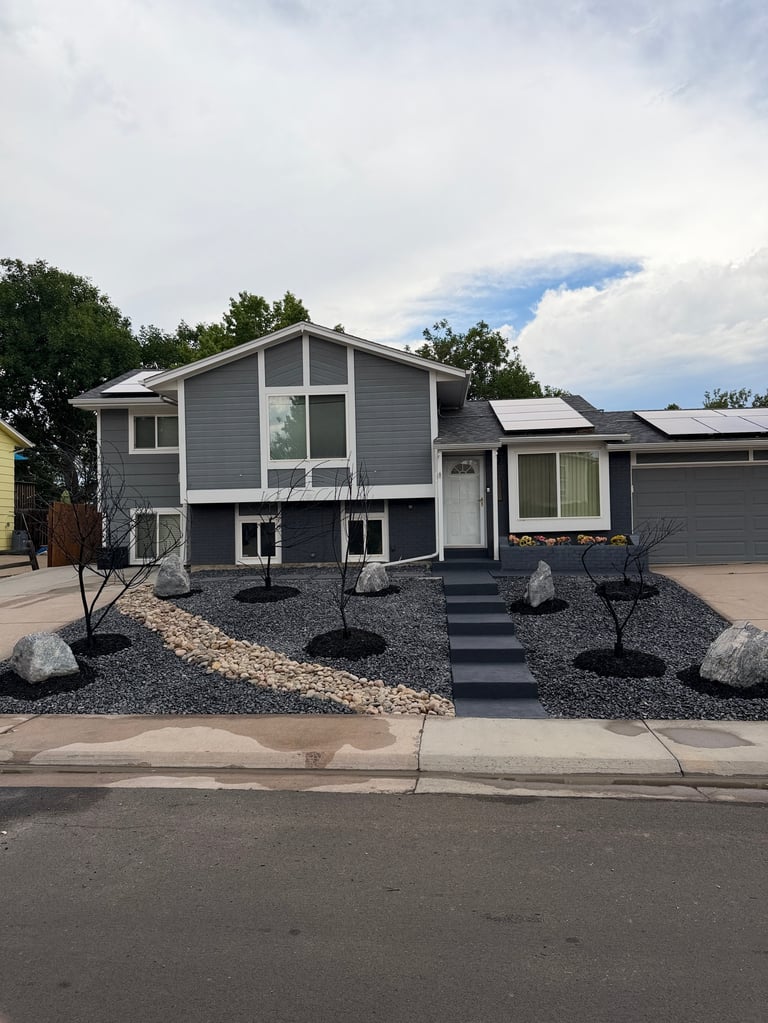 Modern gray house with elevated foundation, large windows, and drought-resistant landscaping with rocks and young trees