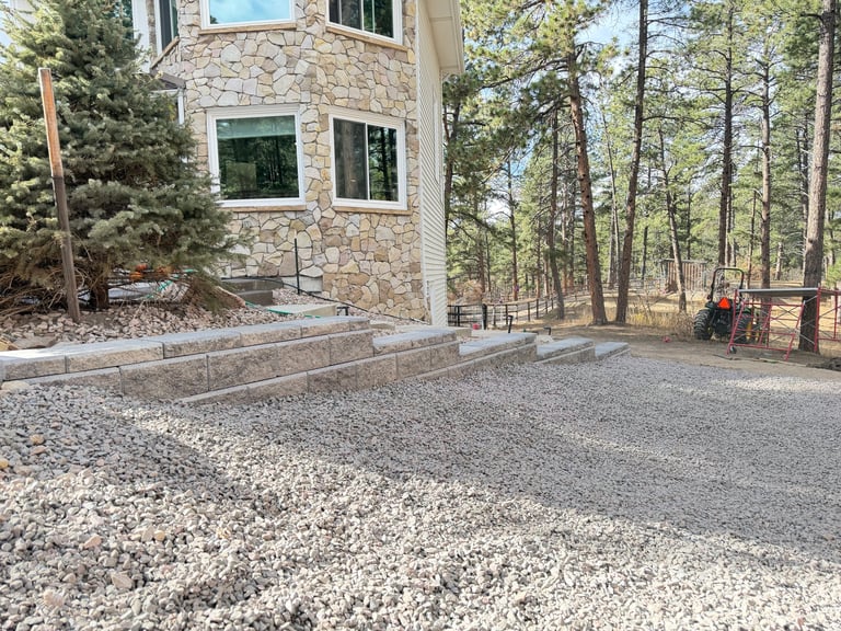 Modern stone house with gravel driveway surrounded by tall pine trees and wooden fence